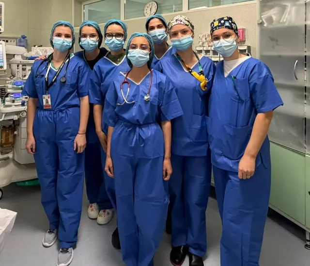Șapte persoane purtând uniforme medicale albastre și măști de protecție stau grupate într-o încăpere cu echipamente medicale și dulapuri metalice.
