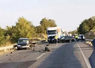 Un autoturism negru și un camion alb sunt pe un drum asfaltat lângă resturi de vehicule și persoane purtând veste reflectorizante.
