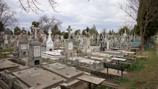 Un cimitir cu numeroase morminte de piatră, cruci și câteva bănci de lemn sub un cer înnorat.