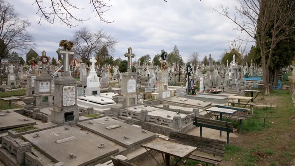 Un cimitir cu numeroase morminte de piatră, cruci și câteva bănci de lemn sub un cer înnorat.
