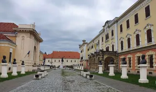 O stradă pavată cu pietriș se întinde între clădiri istorice galbene și albe, având busturi pe piedestale albe dispuse pe margini.