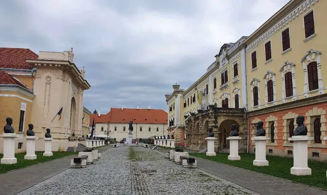 O stradă pavată cu pietriș se întinde între clădiri istorice galbene și albe, având busturi pe piedestale albe dispuse pe margini.