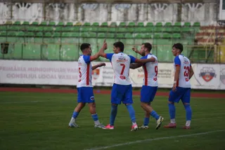 Cinci jucători de fotbal în echipamente alb-roșii și blu sunt pe un teren de fotbal verde în fața unor tribune cu scaune verzi.