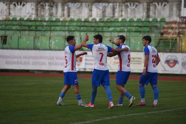 Cinci jucători de fotbal în echipamente alb-roșii și blu sunt pe un teren de fotbal verde în fața unor tribune cu scaune verzi.