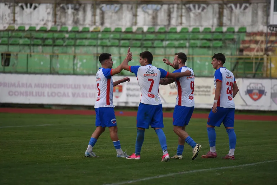 Cinci jucători de fotbal în echipamente alb-roșii și blu sunt pe un teren de fotbal verde în fața unor tribune cu scaune verzi.