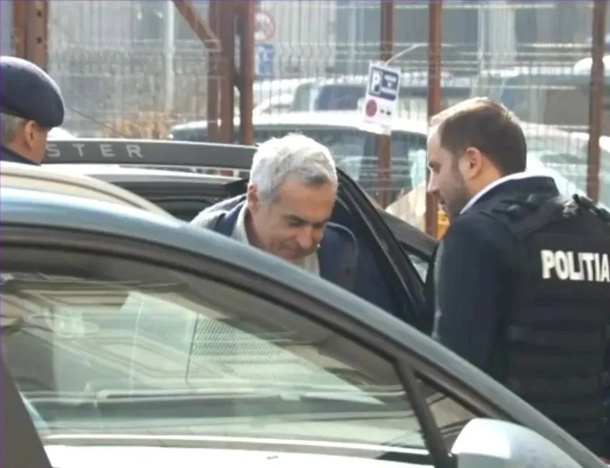 Un bărbat cu părul cărunt este așezat într-o mașină, lângă un agent care poartă un vest cu inscripția POLITIA.