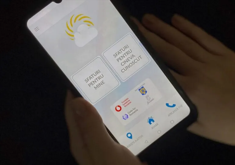 O persoană ține un smartphone care afișează o interfață cu text în limba română și diverse pictograme pe un fundal gri.