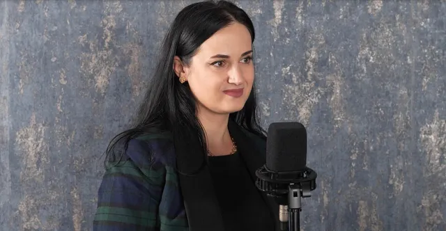 O femeie cu părul negru lung, purtând un sacou în culori închise, stă lângă un microfon negru în fața unui fundal texturat gri.