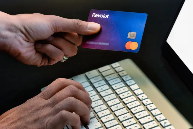 O persoană ține un card Revolut deasupra unui laptop în timp ce degetele celelalte tastrează pe o tastatură de culoare deschisă.