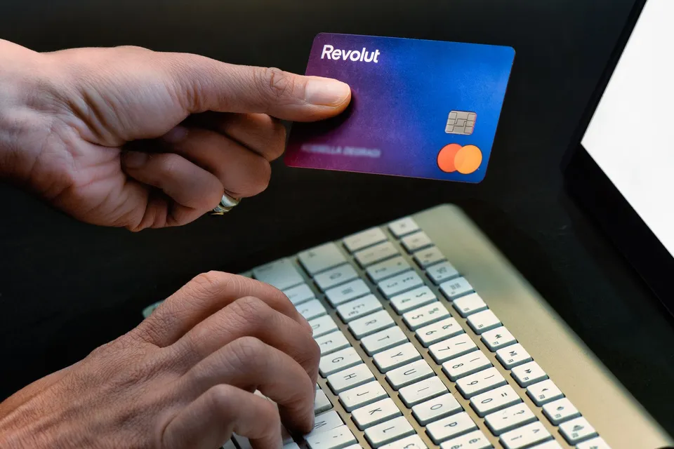 O persoană ține un card Revolut deasupra unui laptop în timp ce degetele celelalte tastrează pe o tastatură de culoare deschisă.