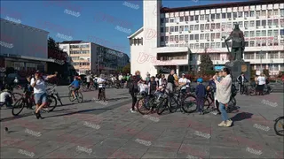 Un grup de oameni stau pe o piață pavată lângă biciclete, în fața unei clădiri mari și a unei statui.