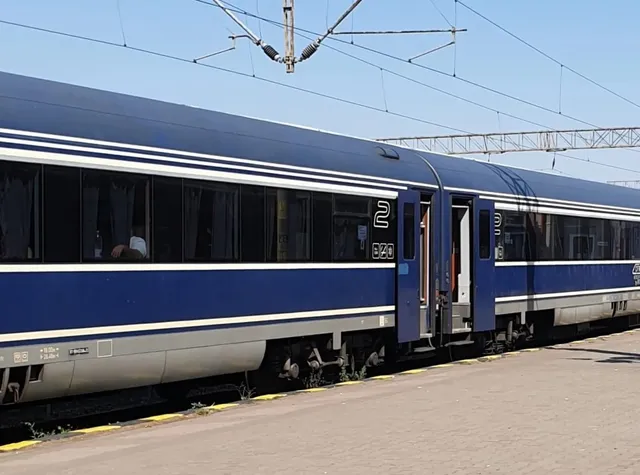Un tren albastru cu ferestre negre și dungi albe este oprit pe un drum de beton sub cabluri electrice de susținere.