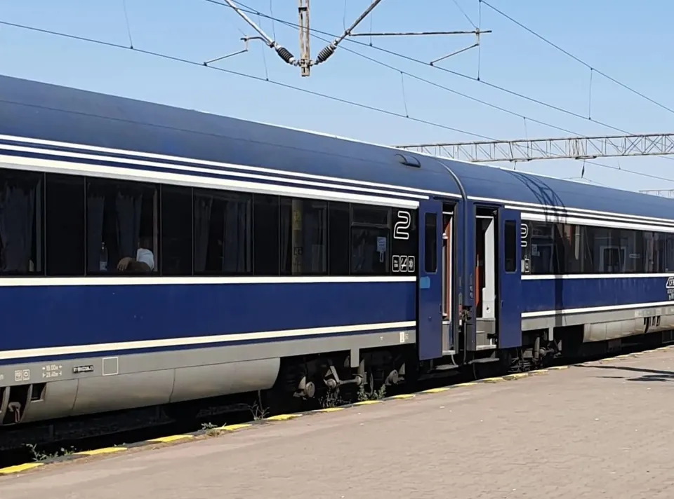 Un tren albastru cu ferestre negre și dungi albe este oprit pe un drum de beton sub cabluri electrice de susținere.
