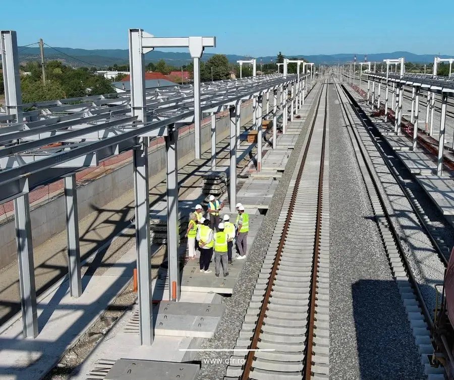 O vedere aeriană a unei șine de cale ferată nou construite, cu structuri metalice pentru electrificare și un grup de muncitori în veste reflectorizante pe peron.