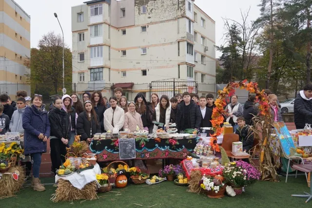 Un grup de persoane stă în spatele unor mese acoperite cu fețe de masă colorate, pe care sunt așezate diverse fructe, flori și decorațiuni de toamnă.