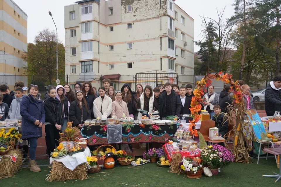 Un grup de persoane stă în spatele unor mese acoperite cu fețe de masă colorate, pe care sunt așezate diverse fructe, flori și decorațiuni de toamnă.