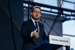 Un bărbat cu ochelari și costum închis la culoare vorbește de la un podium alb pe care scrie un text în limba română.