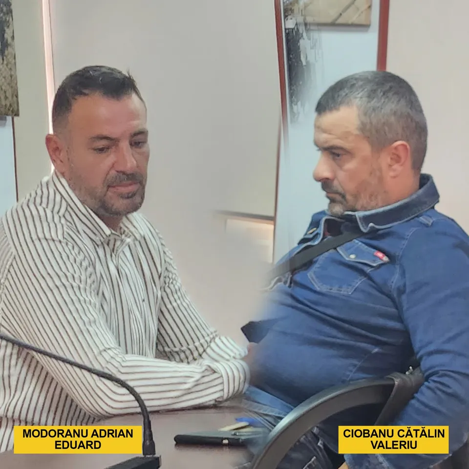 Un bărbat cu cămașă în dungi și un bărbat cu cămașă albastră stau la o masă într-o încăpere.