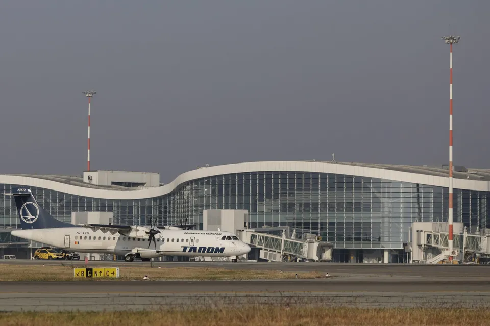 Un avion alb cu marcaje albastre și verzi stă pe pista de lângă un terminal de aeroport cu fațadă de sticlă și două turnuri înalte.