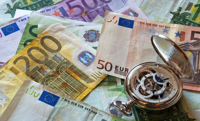 Un ceas de buzunar deschis este așezat pe mai multe bancnote de euro împrăștiate pe o suprafață plană.