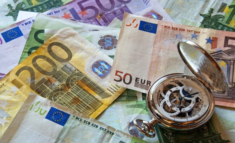 Un ceas de buzunar deschis este așezat pe mai multe bancnote de euro împrăștiate pe o suprafață plană.