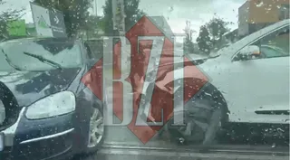 Două autoturisme, unul de culoare închisă și unul alb, sunt poziționate în contact pe un drum sub un cer înnorat.
