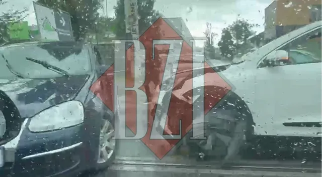 Două autoturisme, unul de culoare închisă și unul alb, sunt poziționate în contact pe un drum sub un cer înnorat.