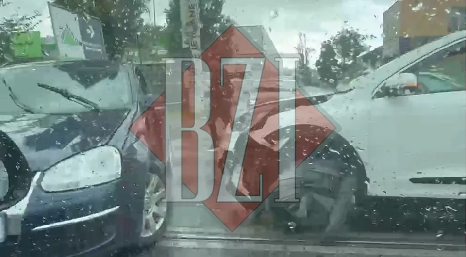 Două autoturisme, unul de culoare închisă și unul alb, sunt poziționate în contact pe un drum sub un cer înnorat.