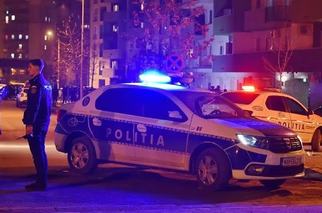 O mașină de poliție albă cu lumini albastre aprinse este parcată pe un drum, lângă un polițist care stă în picioare.