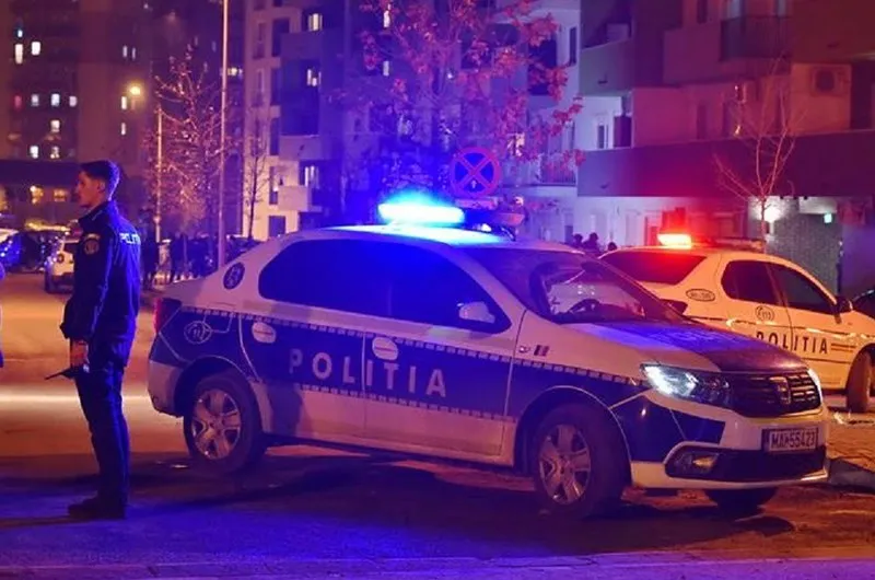 O mașină de poliție albă cu lumini albastre aprinse este parcată pe un drum, lângă un polițist care stă în picioare.