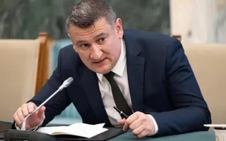 Un bărbat într-un costum închis la culoare și cravată neagră stă la o masă cu un microfon și un caiet deschis în față.