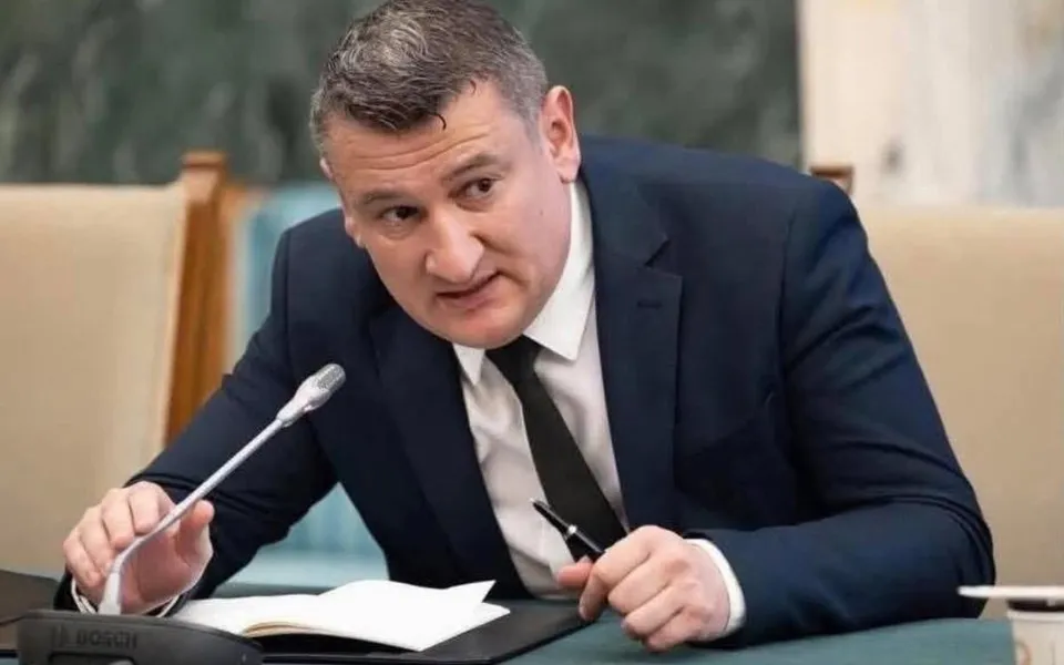 Un bărbat într-un costum închis la culoare și cravată neagră stă la o masă cu un microfon și un caiet deschis în față.