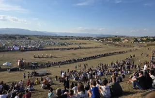 O mulțime de oameni stă pe un câmp deschis sub un cer senin, privind spre o zonă de spectacol delimitată de linii de oameni.