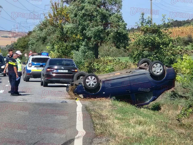 Un autoturism albastru este răsturnat pe o parte într-un șanț, lângă un drum asfaltat unde sunt parcate alte două mașini și stau trei persoane.