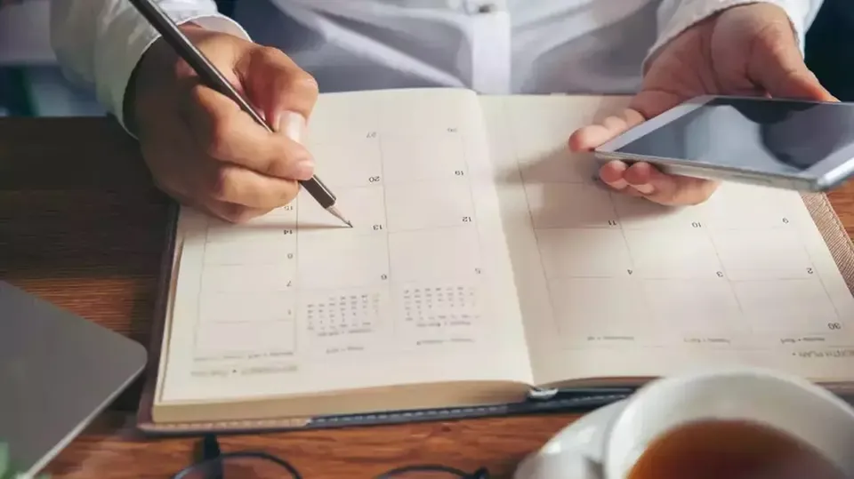 Persoană la birou notează într-un calendar deschis, în timp ce ține un telefon; în prim-plan apar o cană și ochelari.