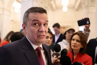 Un bărbat cu părul scurt și costum închis cu cravată roșie privește spre lateral în timp ce este înconjurat de alte persoane și microfoane.