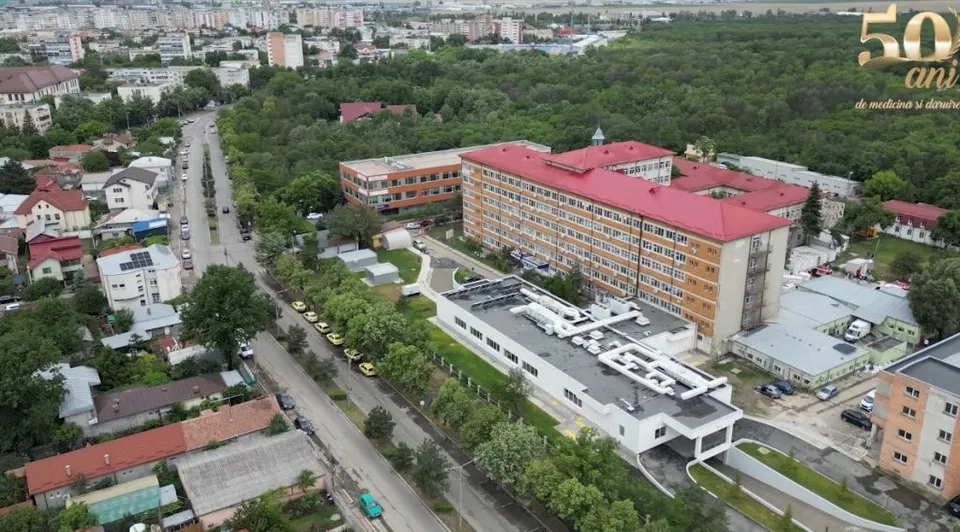 O vedere aeriană prezintă un complex de clădiri cu acoperișuri roșii și albe, înconjurate de copaci verzi și străzi cu câteva mașini parcate.
