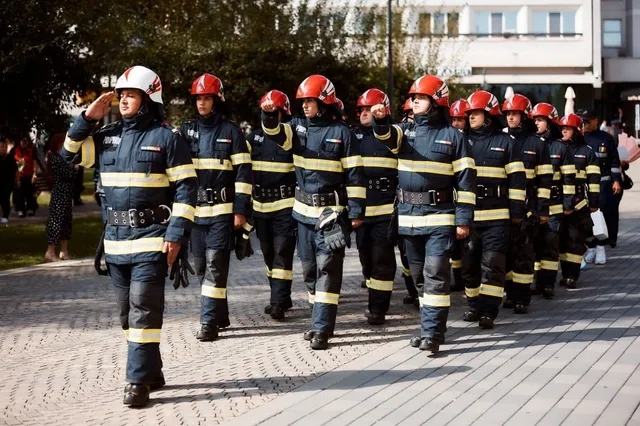 Un grup de pompieri, îmbrăcați în uniforme de intervenție cu dungi reflectorizante și căști, defilează pe o alee pavată, în timpul zilei.