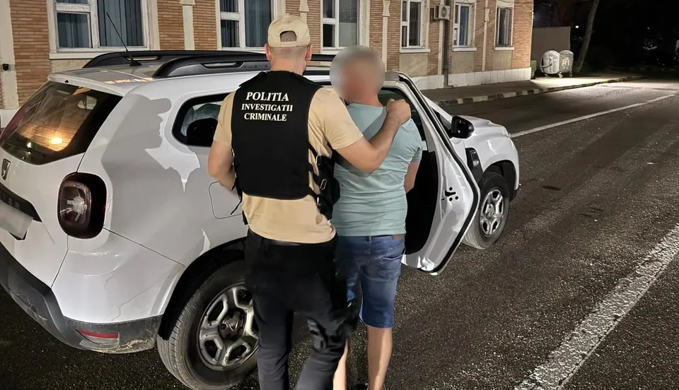 Un agent de poliție în vestă neagră ține un bărbat cu tricou albastru și pantaloni scurți lângă o mașină albă pe un drum noaptea.