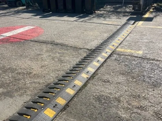 Un dispozitiv de tip bumper din cauciuc negru cu marcaje galbene este montat pe o suprafață asfaltată lângă o marcaj rutier roșu și alb.