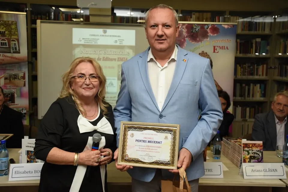 Un bărbat într-un sacou albastru și o femeie într-o bluză neagră cu detalii albe stau lângă o masă cu cărți și un certificat.