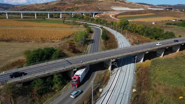 O pod de beton traversează o șosea și o linie feroviară curbată într-un peisaj rural cu câmpuri și dealuri sub un cer deschis.