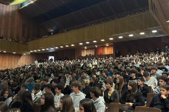 O mulțime de tineri stă așezată pe scaune în interiorul unui auditorium mare cu balustrade de lemn și lumină artificială.