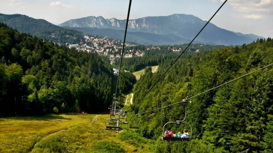 Un telescaun cu oameni transportați trece deasupra unei păduri de conifere și a unei pajiști verzi, sub un cer înconjurat de munți.