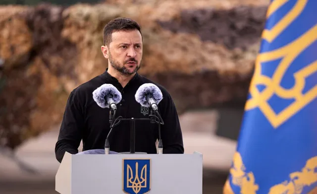 Un bărbat cu barbă și haină neagră stă în spatele unui podium gri cu un simbol galben, având două microfoane în față.