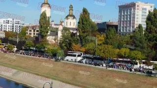 O linie de autobuze albe și negre este parcată lângă un gard, în fața unor copaci și clădiri sub un cer senin.