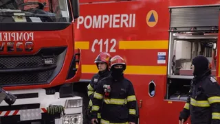 Trei pompieri în echipament de protecție negru cu benzi reflectorizante galbene stau lângă un autospecială de pompieri de culoare roșie.