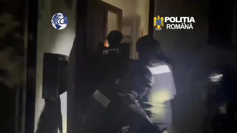 Patru persoane în uniformă întunecată stau într-un spațiu slab iluminat lângă un perete și o ușă deschisă.