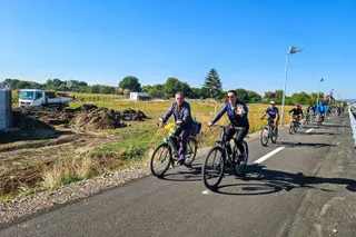 Mai mulți bicicliști parcurg o cale asfaltată într-un peisaj însorit, având un teren accidentat și un camion alb în fundal.