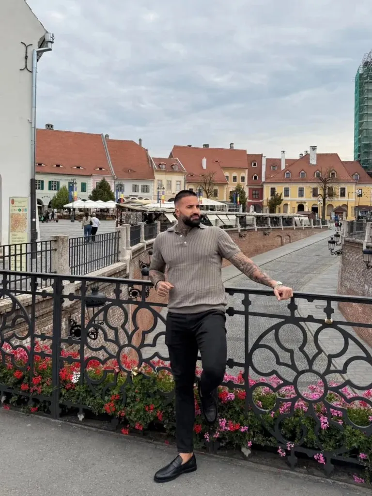 Un bărbat cu tatuaje poartă un tricou gri și pantaloni negri, stând lângă un gard metalic decorat cu flori roz și roșii.